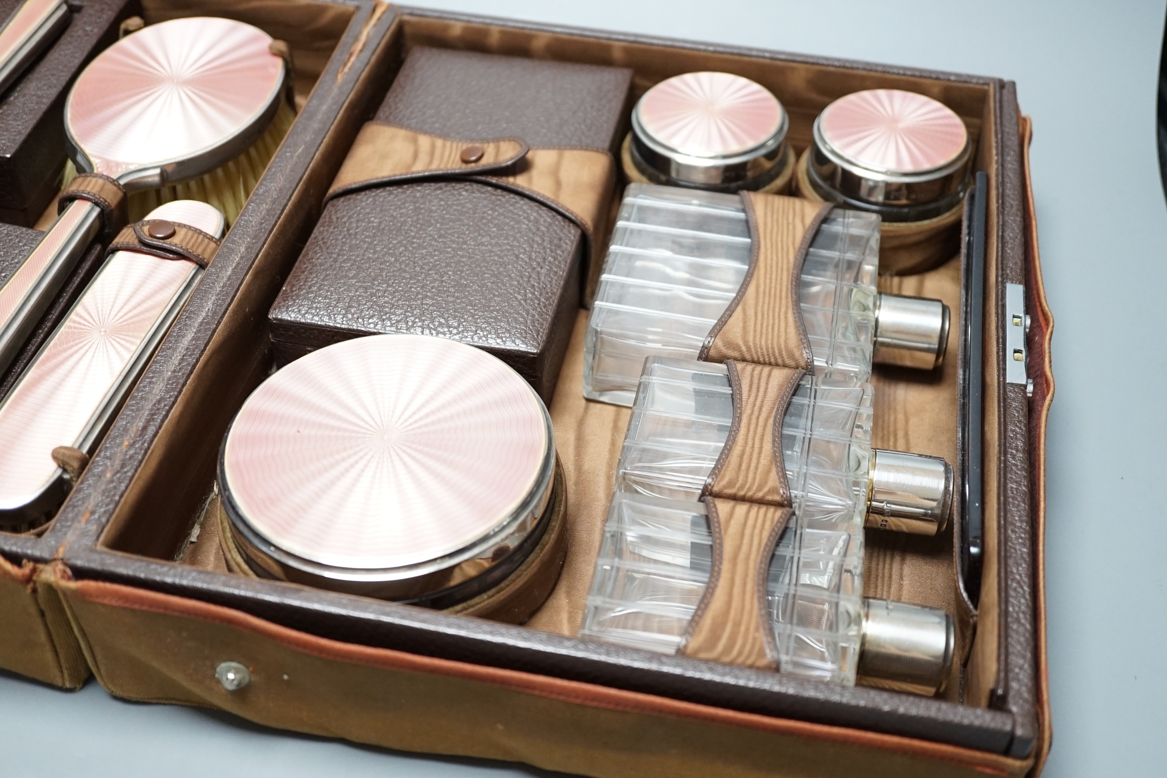 A George V travelling toilet case, containing eleven silver and pink enamel mounted glass bottles jars, mirror and brushes, Mappin & Webb, 1936, in brown leather case, case width 31cm.